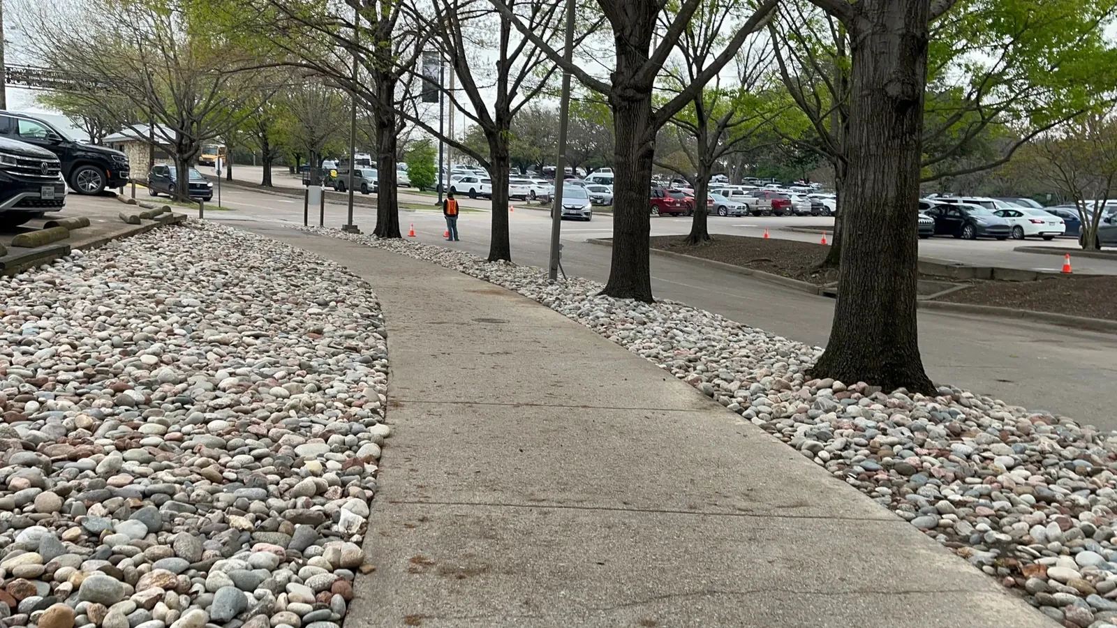 River rock beds flanking a walkway on a commercial parking campus, with a crew member working a coned-off zone beyond