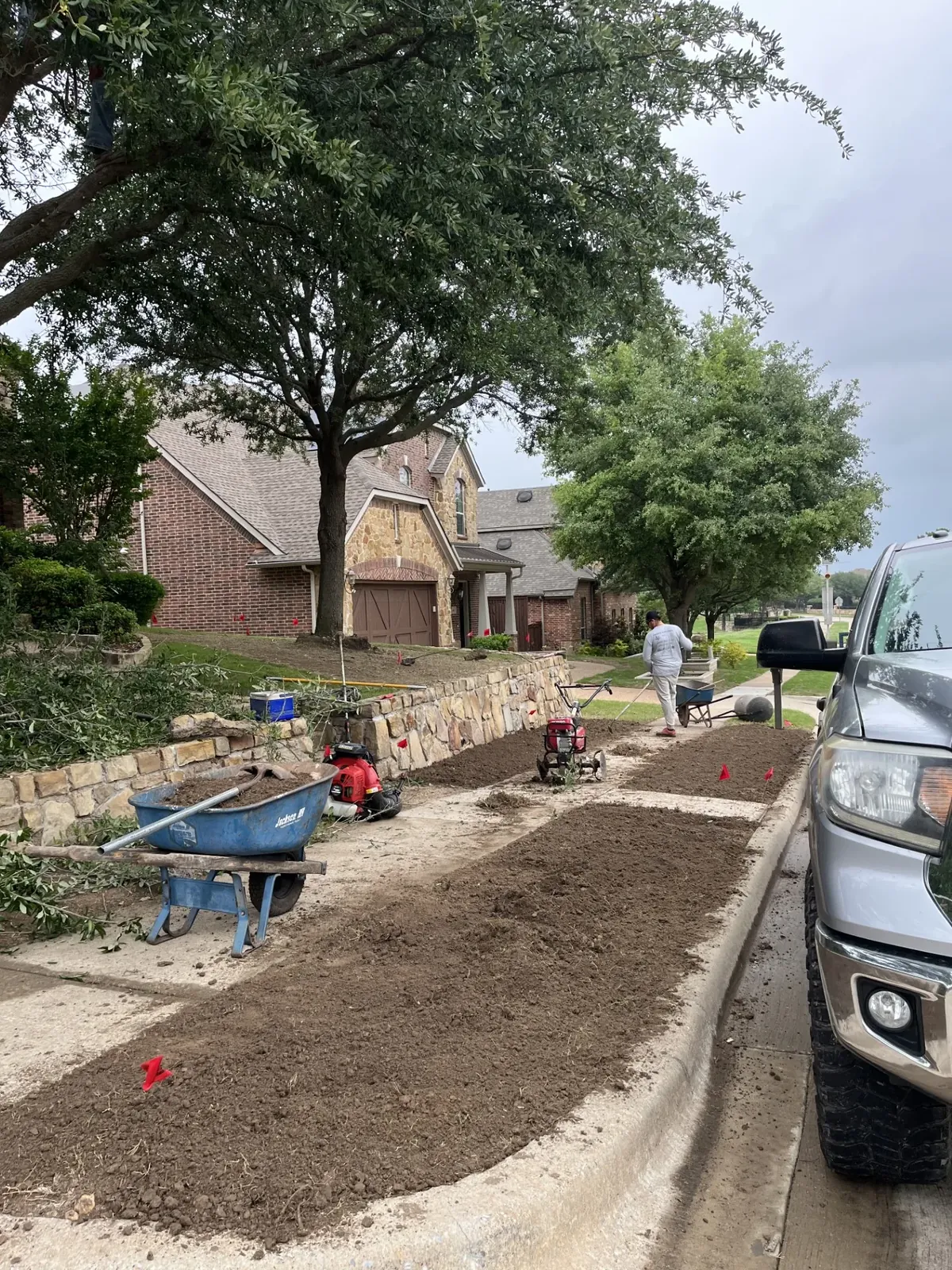 Crew tilling bare dirt on tiered front yard with stone retaining wall before sod installation, North Texas