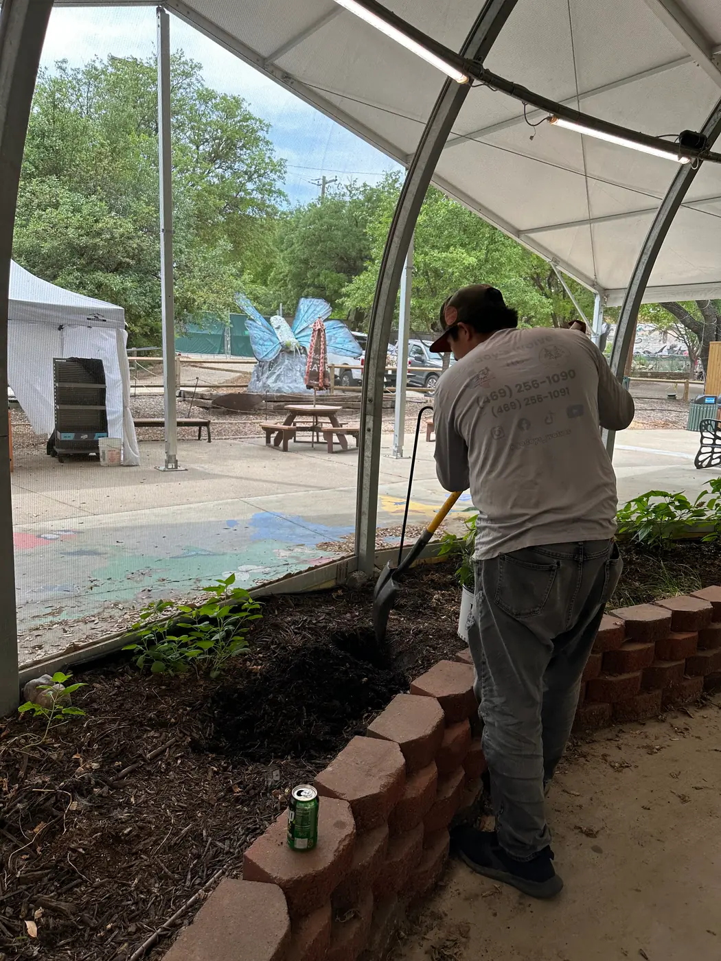 Godoy's Wolves crew member turning soil in a raised brick bed at the butterfly house, with the giant blue butterfly sculpture visible outside