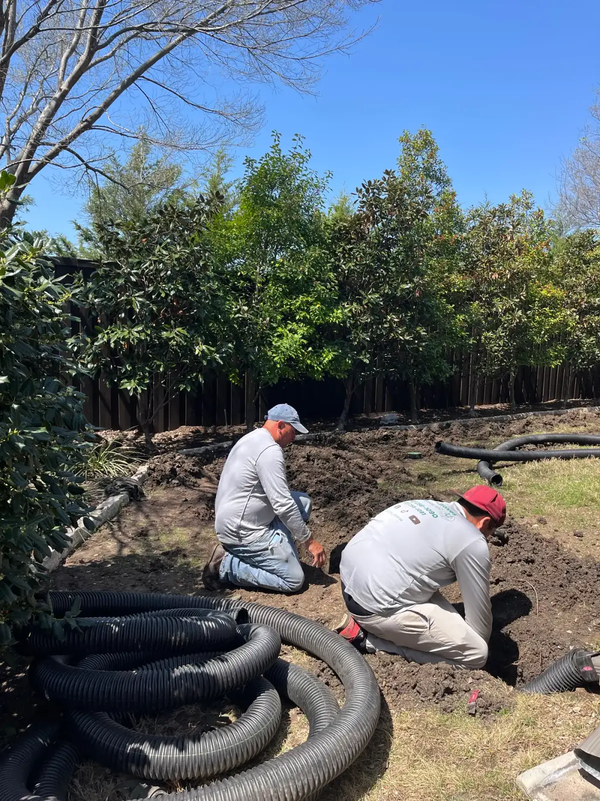 Two Godoy's Wolves crew members setting perforated drain pipe into an open trench along a fence line
