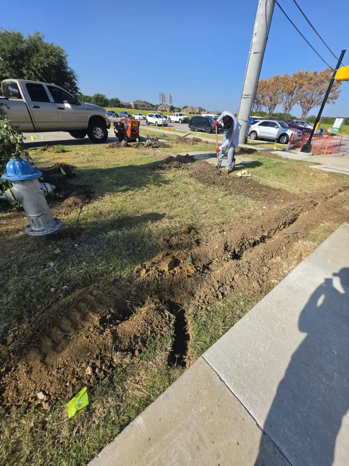 Godoy's Wolves crew member digging an irrigation trench along a commercial frontage with a trencher and utility locate flags on site