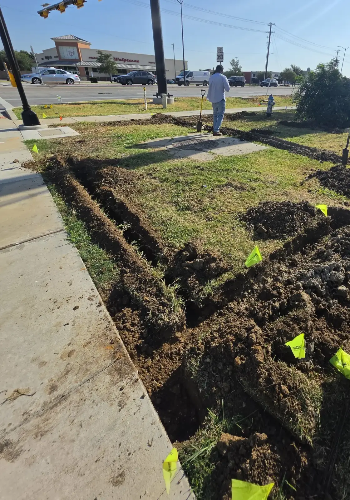 Irrigation trench run opened along a commercial frontage with utility locate flags marking the line
