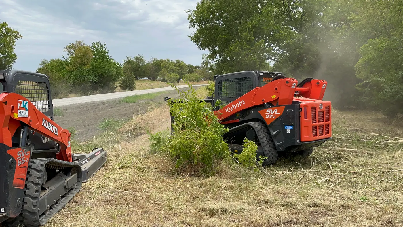 Two tracked skid steers with brush-cutter attachments clearing heavy brush along a rural fence line, cut material windrowed behind them