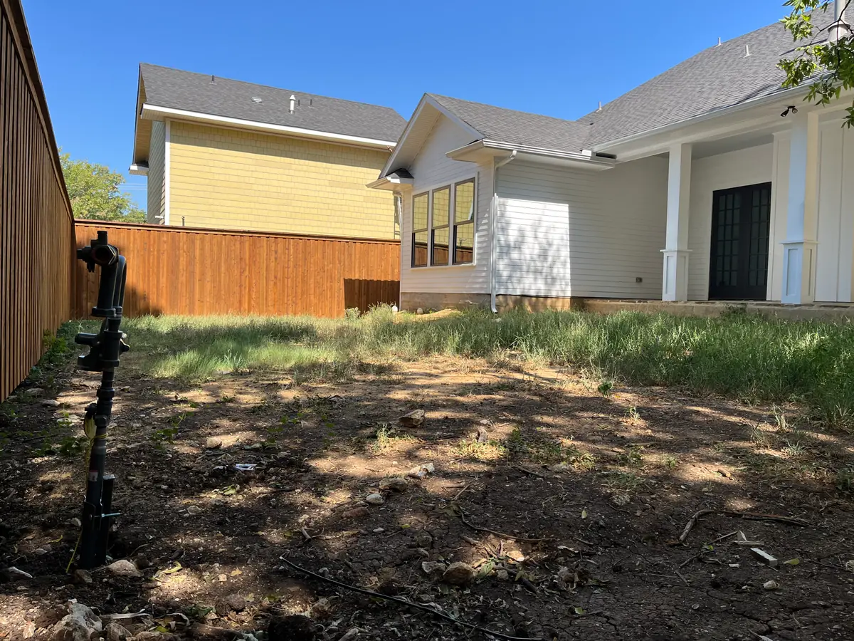 Back yard of a new build before landscaping: cracked bare ground and weeds against a new cedar fence, with a backflow preventer riser standing at the property line