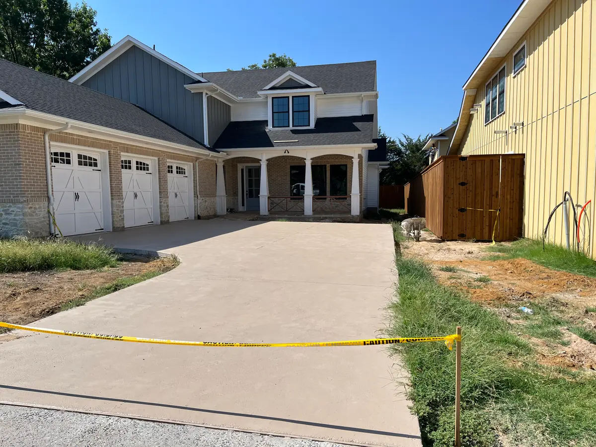 New brick home with three garage doors and a freshly poured driveway still taped off, the lot either side still bare dirt and weeds