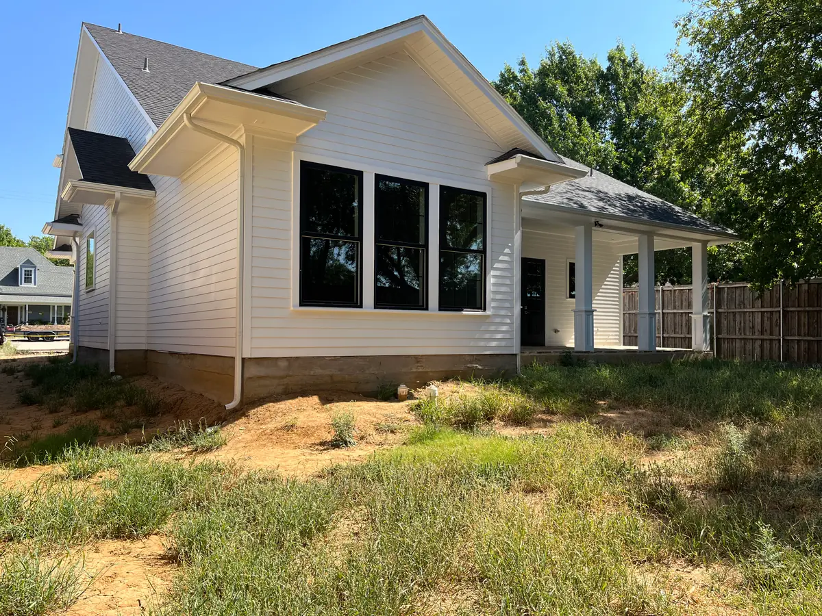 White board-and-batten farmhouse elevation on a new build, the yard in front of it rough weeds and exposed dirt with irrigation stub-outs showing at the slab