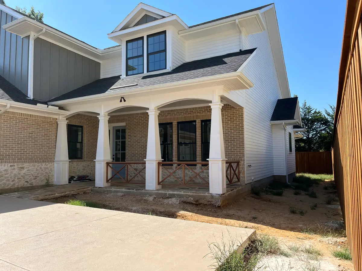Covered brick porch with white columns and a cedar X-brace rail on a new build, the ground in front of it bare dirt and scattered weeds with no beds cut yet