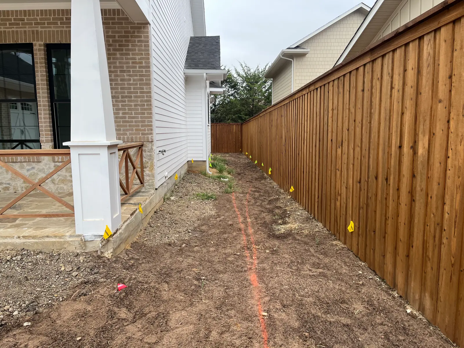 Side yard on a new build graded flat and dressed, an orange layout line sprayed down the run and yellow utility locate flags set along a new cedar fence
