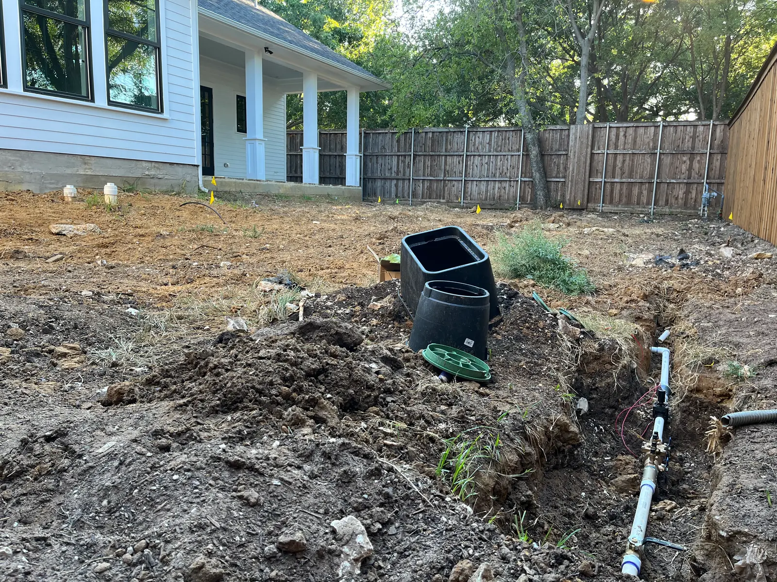 Irrigation valve manifold wired and set in an open trench with valve boxes and marking flags beside it, a new white farmhouse behind