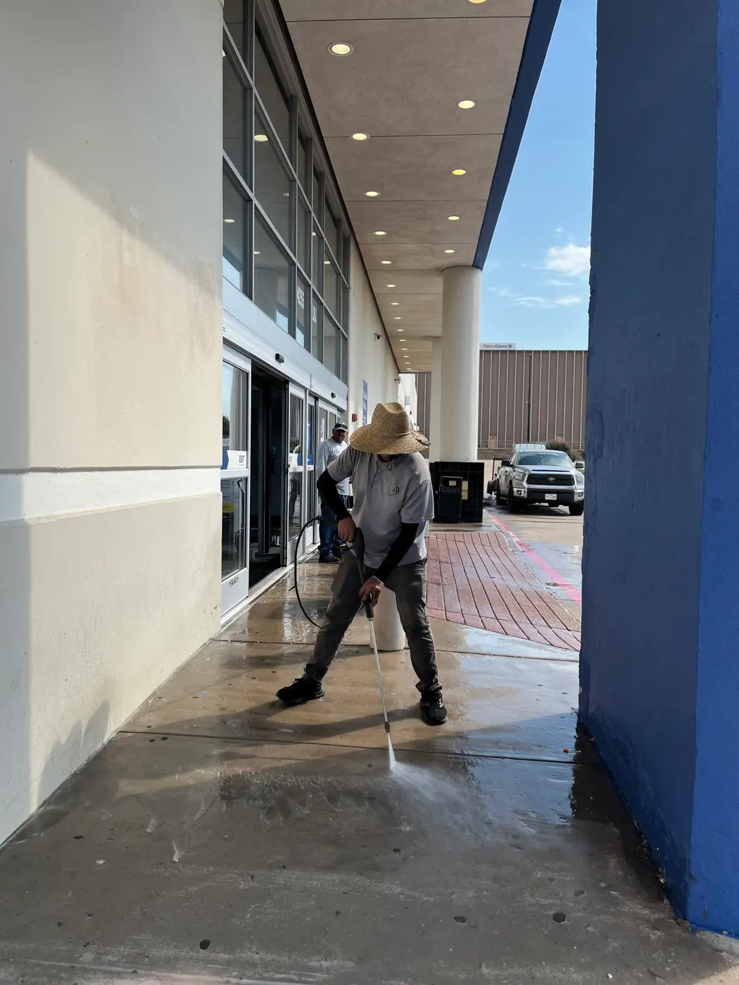 Crew member pressure washing a commercial building walkway, spray visible at the wand